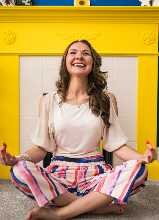 Kat sits cross legged and looks up towards the ceiling smiling. 